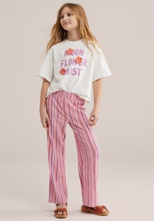 Meisje staand met ontspannen handen, draagt een wit T-shirt met bloemapplicaties en lavendelroze gestreepte broek en bruine sandalen.