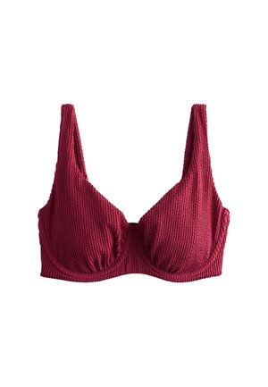 Žena v červenom bikinách stojaca vonku, usmievajúca sa, obklopená zelenými rastlinami a rozmazanými ružovými kvetmi v pozadí.