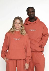 Două modele îmbrăcate în hanorace și pantaloni de culoare ruginie, cu logo-ul "MULTIPLY APPAREL", stând pe un fundal alb simplu.