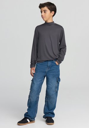 Koszula z długim rękawem w ciemnoszarym kolorze z wysokim kołnierzem, zestawiona z luźnymi, niebieskimi spodniami typu cargo z licznymi kieszeniami oraz czarnymi trampkami.