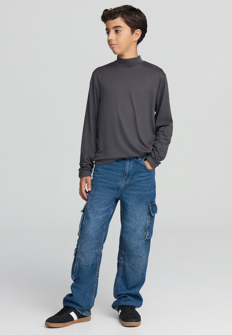 Koszula z długim rękawem w ciemnoszarym kolorze z wysokim kołnierzem, zestawiona z luźnymi, niebieskimi spodniami typu cargo z licznymi kieszeniami oraz czarnymi trampkami.