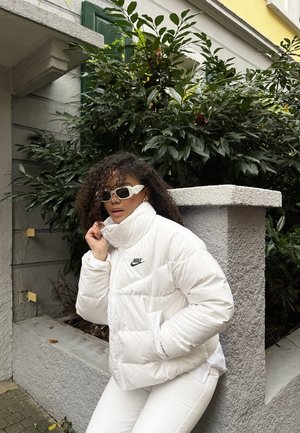 Doudoune blanche avec col montant, arborant un logo noir de Nike. Associée à un pantalon blanc et des lunettes de soleil blanches distinctives. Fond en mur gris.