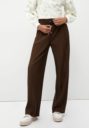 Osoba ubrana w brązowe szerokie spodnie z pionowymi plisami i elastycznym pasem, nosząca białe sneakersy, regulująca sznurek w talii.