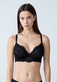 Sostén de encaje negro con bordes festoneados, soporte de aros y tirantes ajustables. El diseño de encaje presenta patrones y texturas florales.