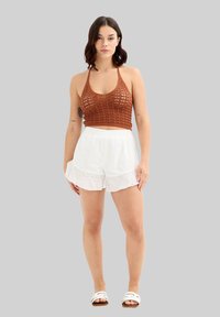 Top de ganchillo marrón con tirantes, combinado con pantalones cortos blancos, con volantes y ojales. El conjunto presenta un ajuste relajado y detalles de tela texturizada.