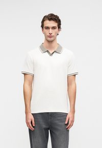 Junger Mann steht und trägt ein weißes kurzärmliges Poloshirt mit grauem Kragen und grauen Jeans vor einem schlichten hellen Hintergrund.