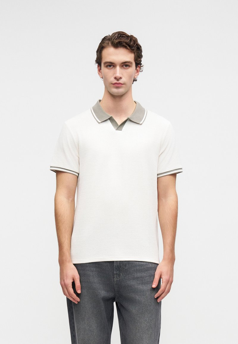 Junger Mann steht und trägt ein weißes kurzärmliges Poloshirt mit grauem Kragen und grauen Jeans vor einem schlichten hellen Hintergrund.