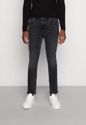 Czarne obcisłe jeansy noszone z białymi trampkami i czarną prążkowaną bluzką z długim rękawem, stojące na jednolitym jasnym tle.