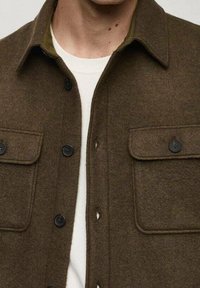 Brązowa koszula dżinsowa z mieszanki wełny, z klasycznym kołnierzem, dwiema kieszeniami na piersi i czarnymi guzikami, w połączeniu z białą koszulką pod spodem.