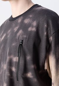 Persona con una camiseta de teñido anudado en negro y beige, que tiene un bolsillo con cremallera vertical en el pecho y un cuello negro.