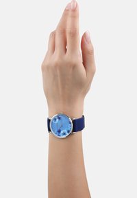 Rond horloge met een blauwe bloemenmotief wijzerplaat, zilveren bezel en een blauwe siliconen band, gedragen aan een hand met een neutrale huidtint.
