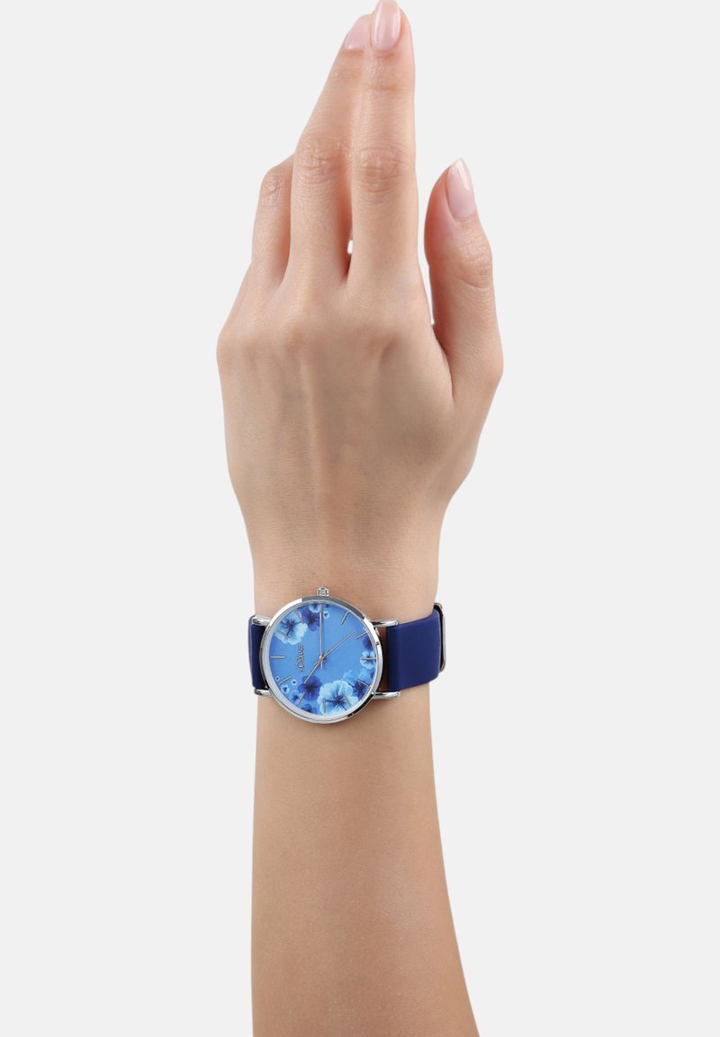 Rond horloge met een blauwe bloemenmotief wijzerplaat, zilveren bezel en een blauwe siliconen band, gedragen aan een hand met een neutrale huidtint.