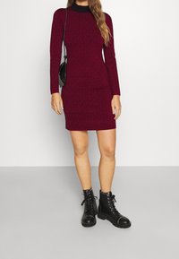 Robe en maille bordeaux à manches longues, col montant, avec un motif texturé. Portée avec des bottes de combat noires à lacets ornées de clous argentés.