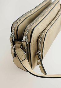 Beige rektangulær skuldertaske med to sølvlynlåse, sort kantning, dobbelte rum og en fastgjort justerbar rem.