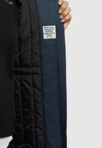 Das Innere der schwarz gefütterten Jacke zeigt einen dunkelblauen Außenstoff. Ein Etikett mit Text ist sichtbar. Sie verfügt über eine gesteppte Polsterung und eine glatte Textur.
