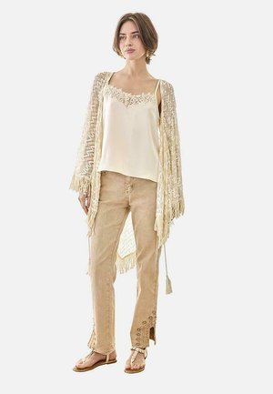 Mujer con chal de encaje color crema, camisola de seda, pantalones beige bordados y sandalias, de pie sobre un fondo blanco.