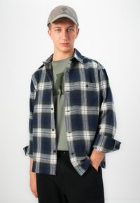 Young man wearing gray cap, green graphic tee, open blue and white plaid shirt with rolled sleeves, and black pants, posing with one hand in pocket.