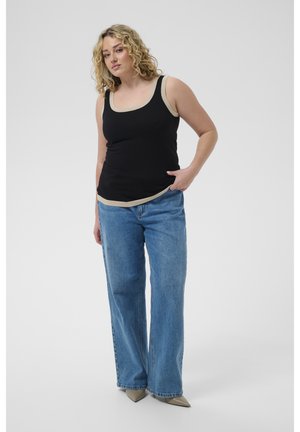 Femme aux cheveux blonds bouclés portant un haut noir sans manches avec un bord beige, un jean large bleu et des chaussures beiges pointues, debout, la main dans la poche.