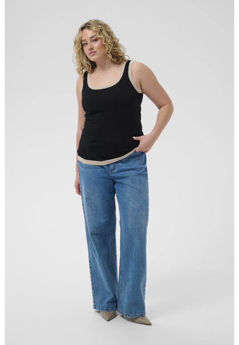Femme aux cheveux blonds bouclés portant un haut noir sans manches avec un bord beige, un jean large bleu et des chaussures beiges pointues, debout, la main dans la poche.