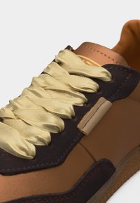 Κοντινή λήψη καφέ αθλητικού παπουτσιού με σκούρο περίγραμμα και λαμπερές κρεμ σατέν κορδόνια που περνούν μέσα από οπές, τονίζοντας τις υφές και τις ραφές.