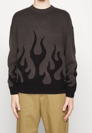 Person wears dark gray knit sweater with black flame pattern and light brown pants, standing against white background.