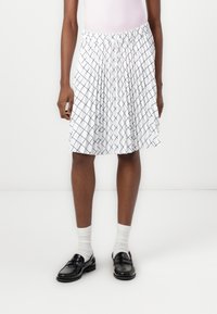 Jupe blanche de longueur genou avec un motif de grille géométrique bleu marine. Elle dispose d'une taille élastiquée et d'un design plissé. Portée avec des mocassins noirs.