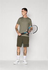 Mann i olivenfarget sportsklær som holder en tennisracket, stående mot en ensfarget lys bakgrunn, med synlige tatoveringer på armer og ben.