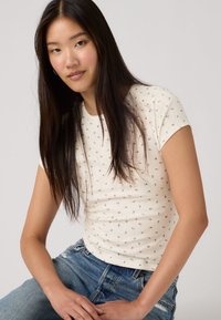 Νεαρή γυναίκα με μακριά σκουρόχρωμα μαλλιά, που φοράει λευκή κοντομάνικη μπλούζα με λουλούδια και μπλε τζιν, καθισμένη μπροστά σε ένα απλό φωτεινό φόντο.