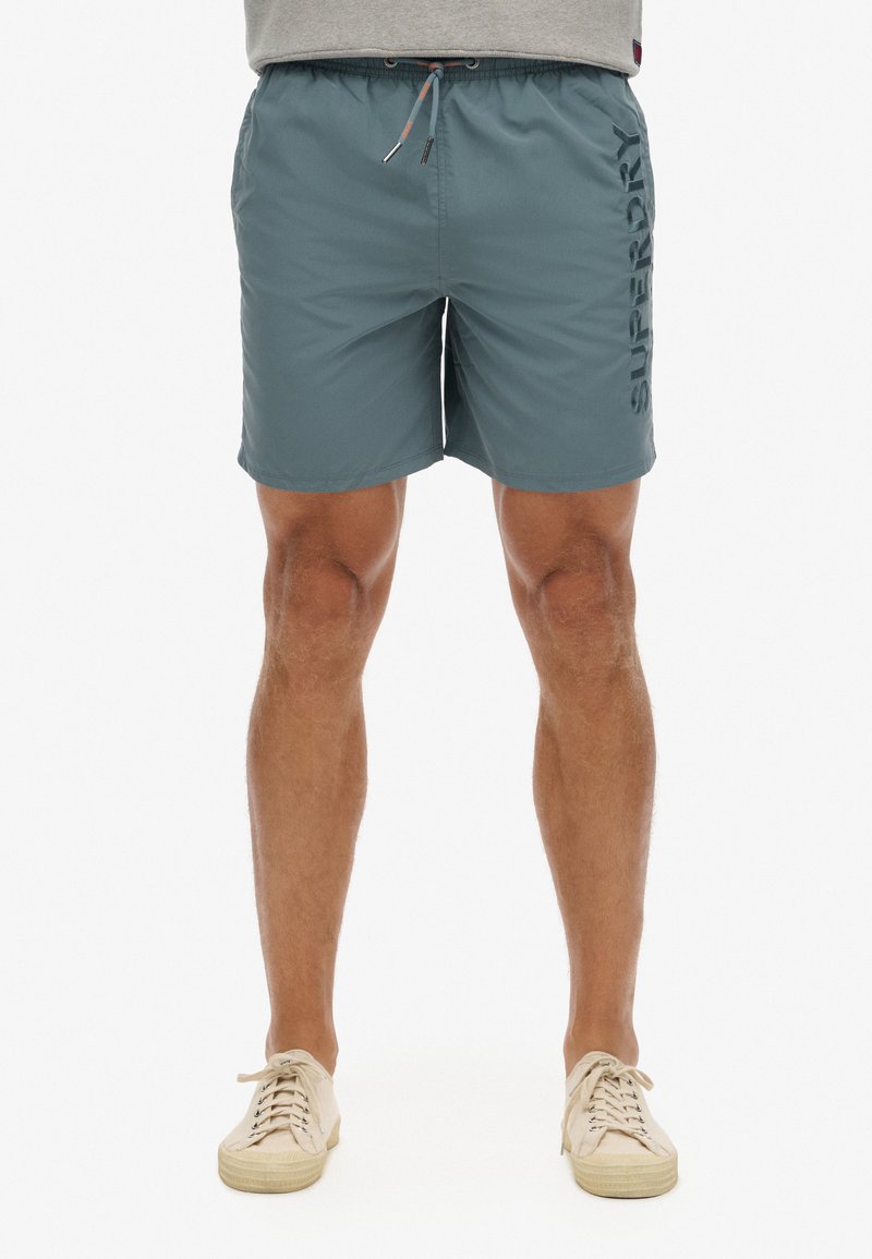 Mannen badshorts in donkergroen met zijzakken, elastische tailleband en trekkoord. Groot "SUPERDRY" logo verticaal opzij gedrukt.