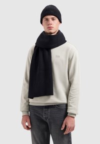 Zwarte gebreide beanie en bijpassende sjaal, gemaakt van textiel met een structuur, gecombineerd met een lichtgrijze sweatshirt en donkere jeans. Simpele, casual outfit.