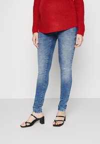 Jeans ajustados de mezclilla azul claro con desgastes en las rodillas. Combinados con un suéter de punto rojo y sandalias negras de tacón con tacón cuadrado.