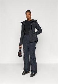Chaqueta negra impermeable con capucha, combinada con pantalones cargo negros. El modelo sostiene un par de gafas. Ambos artículos presentan una textura suave.