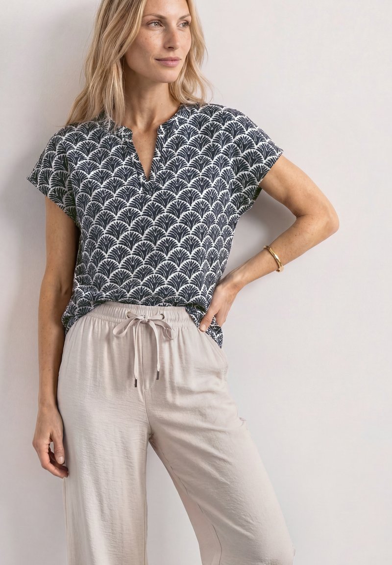 Femme portant un haut à manches courtes à motif noir et blanc et un pantalon beige clair à cordon, debout avec une main sur la hanche devant un fond uni.