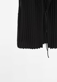 Jupe plissée noire avec un détail de lien à la taille, suspendue sur un fond blanc uni.