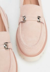 Mocasines de ante en rosa suave con detalles en metal plateado. Suela blanca gruesa, punta redondeada y costuras detalladas alrededor de la parte superior.