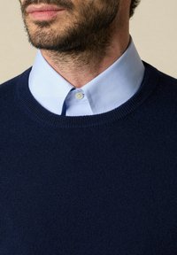 Námořnický pletený svetr s kulatým výstřihem, přes lehkou modrou košilí s límečkem. Hladká textura s viditelným švem na límci.