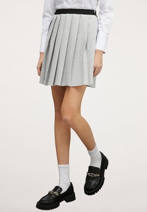 Personne portant une jupe plissée gris clair, une chemise blanche boutonnée, des chaussettes blanches et des mocassins noirs avec un détail de chaîne dorée.