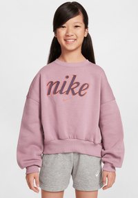 Sudadera de Nike en color lavanda con mangas anchas y dobladillo acanalado, que presenta un logo naranja audaz. Combinada con pantalones cortos deportivos grises.