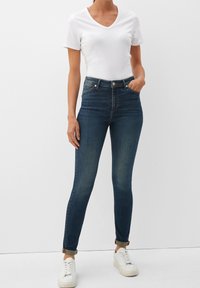 Vaqueros skinny de mezclilla oscura con cintura alta, que presentan una textura suave y un desvanecimiento mínimo, combinados con una camisa blanca de manga corta y escote en V.