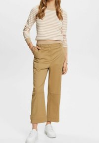 Vrouw in beige wijde broek, beige en witte gestreepte lange mouwen top en witte platform sneakers, staande met één hand in de zak.