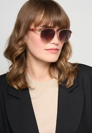 Donna con capelli castano chiaro ondulati che indossa grandi occhiali da sole marroni sfumati, top beige e blazer nero, che guarda leggermente di lato su uno sfondo semplice.