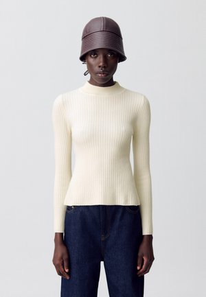 Mujer vestida con un suéter de manga larga blanco de canalé, unos jeans azul oscuro y un sombrero tipo cubo de cuero marrón, de pie contra un fondo liso.