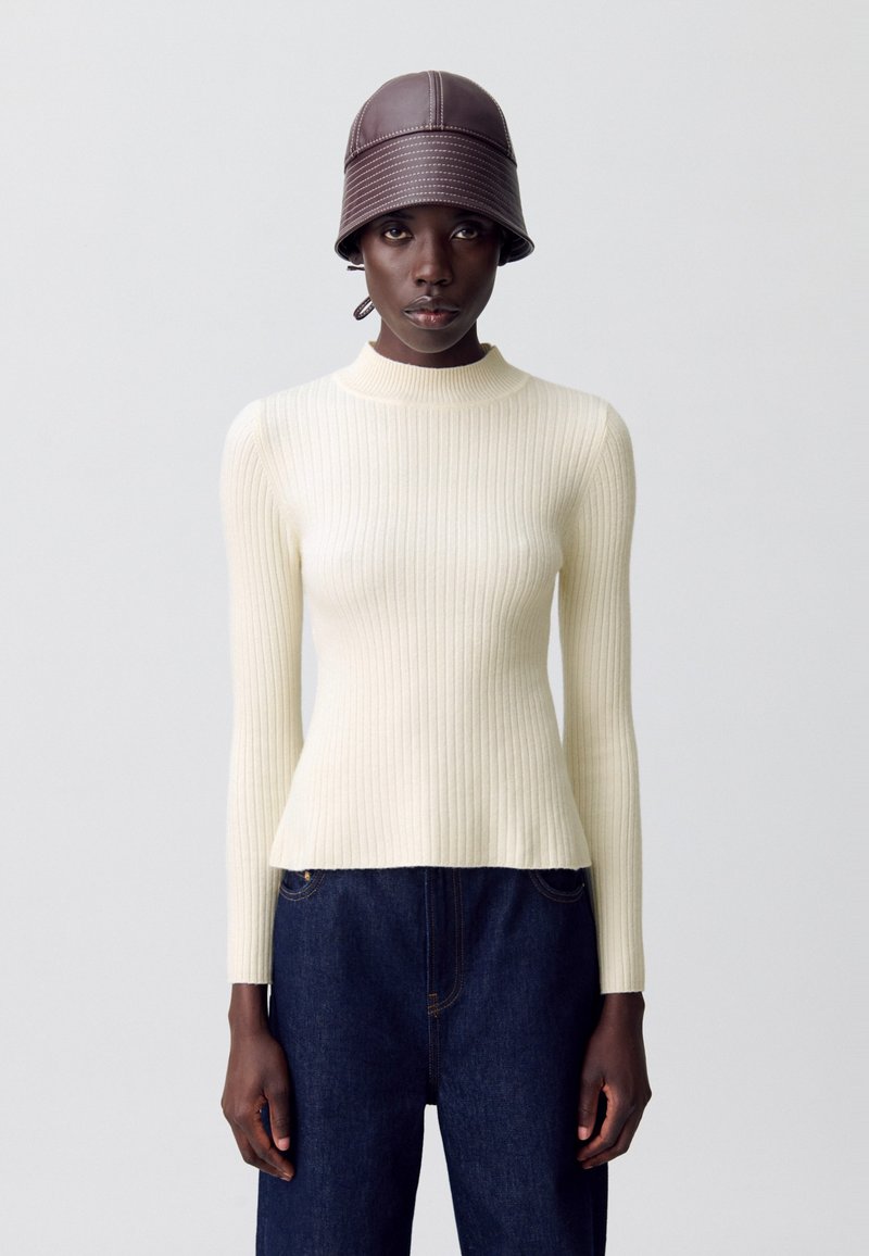 Femme portant un pull à manches longues blanc côtelé, un jean bleu foncé et un chapeau seau en cuir marron, se tenant contre un fond uni.