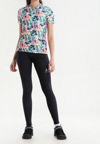 Le haut de sport à manches courtes présente un motif floral multicolore, avec un col zippé quart, associé à des leggings noirs ajustés et des baskets.