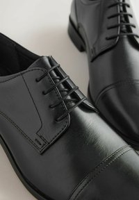 Mustad musta nahast meeste pidulikud jalanõud suletud nööbistamise ja õmmeldud varbakatte detailiga, paigutatud heledale pinnale.