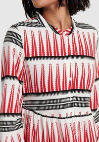 Žena v šatech s dlouhým rukávem, které mají červený, černý a bílý geometrický zigzag vzor a knoflíky po celé přední straně.