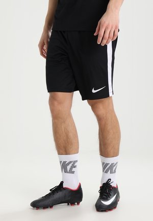 Mężczyzna ubrany w czarne spodenki piłkarskie Nike i czarne korki, z białymi skarpetkami Nike, stoi na białym tle.