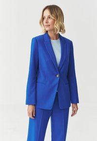 Blue tailored suit featuring a single gold button, notched lapels, and two pockets; paired with a light blue top. Smooth texture.