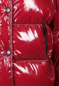 Röd glänsande puffjacka med quiltat mönster, utrustad med en ficka med dragkedja och svarta tryckknappar framtill. Slät, glänsande yta.