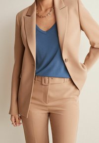 Blazer beige à simple bouton, associé à une chemise bleu en V en dessous. Pantalons beige ajustés avec une ceinture assortie à la taille. Collier en chaîne.